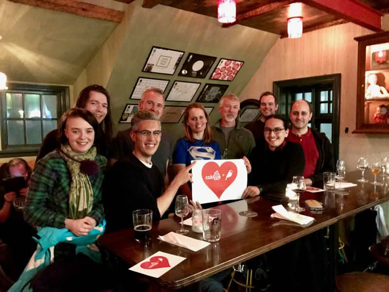 A diverse group of individuals gathered around a table, holding a sign with the Caktus Group and Wagtail CMS logo in a heart shape